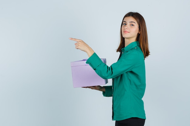 Jovem garota com blusa verde, calça preta segurando uma caixa de presente, apontando para a esquerda e olhando alegre, vista frontal.