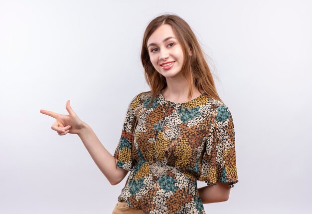 Jovem garota com cabelo comprido usando um vestido colorido parecendo confiante apontando com o dedo indicador para o lado sorrindo