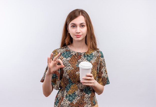 Jovem garota com cabelo comprido usando vestido colorido segurando a xícara de café sorrindo confiante fazendo sinal de ok