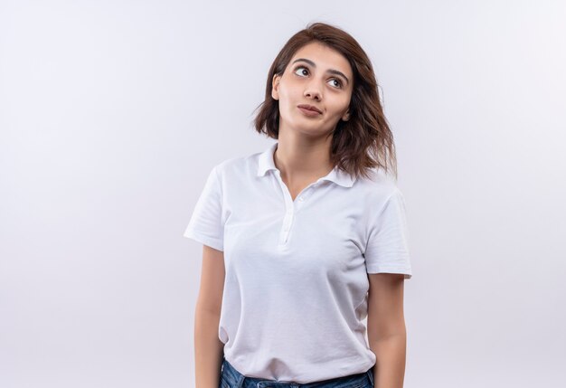 Jovem garota com cabelo curto e camisa pólo branca olhando para o lado com uma expressão triste no rosto