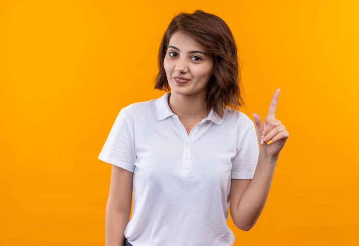 Foto grátis jovem garota com cabelo curto usando uma camisa pólo branca sorrindo confiante apontando com o dedo para cima