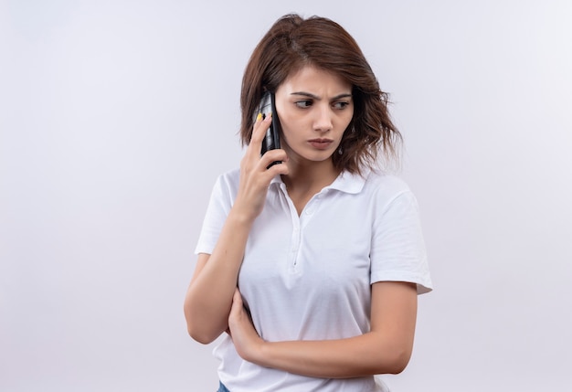 Jovem garota com cabelo curto, vestindo uma camisa pólo branca falando no celular com o rosto carrancudo