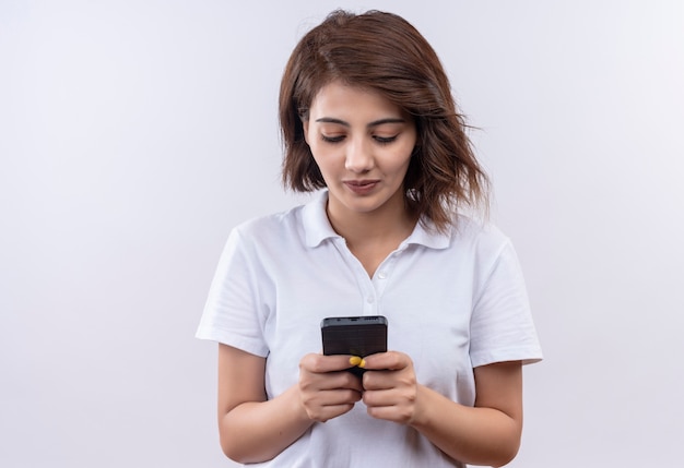 Jovem garota com cabelo curto, vestindo uma camisa pólo branca segurando um telefone celular, conversando com um sorriso no rosto