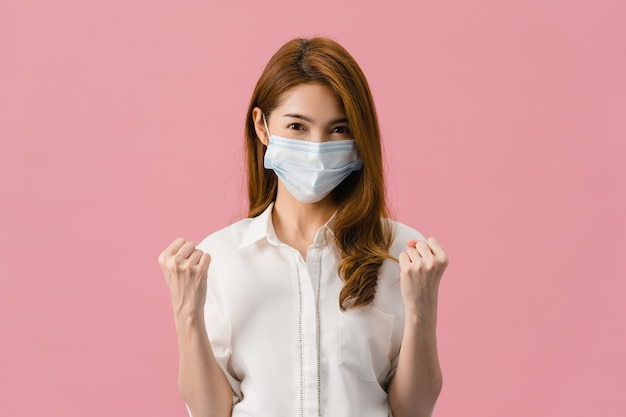 Jovem garota da Ásia usando máscara médica mostrando o símbolo da paz, incentive com vestido de pano casual e olhando para a câmera isolada no fundo rosa.