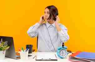 Foto grátis jovem garota de call center usando fone de ouvido, sentada na mesa com ferramentas de trabalho, colocando as mãos no fone de ouvido, olhando para o lado isolado em fundo laranja