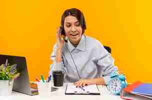 Foto grátis jovem garota do call center impressionada usando headset, sentada na mesa, colocando a mão no headset, olhando para o lado isolado em laranja