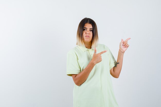 Jovem garota em t-shirt apontando para a direita com o dedo indicador e olhando sério, vista frontal.