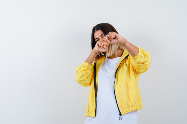 Jovem garota em t-shirt branca, jaqueta amarela cerrando os punhos, em pé em pose de luta e olhando confiante, vista frontal.