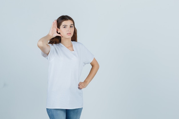 Jovem garota em t-shirt branca ouvindo uma conversa privada e olhando curiosa, vista frontal.