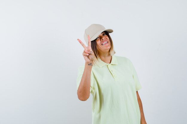 Jovem garota em t-shirt e boné, mostrando o gesto de paz e olhando feliz, vista frontal.