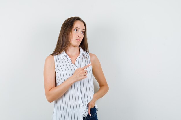 Jovem garota em t-shirt, jeans, apontando para o lado e parecendo impotente, vista frontal.