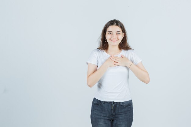 Jovem garota em t-shirt, jeans de mãos dadas no peito e olhando feliz, vista frontal.