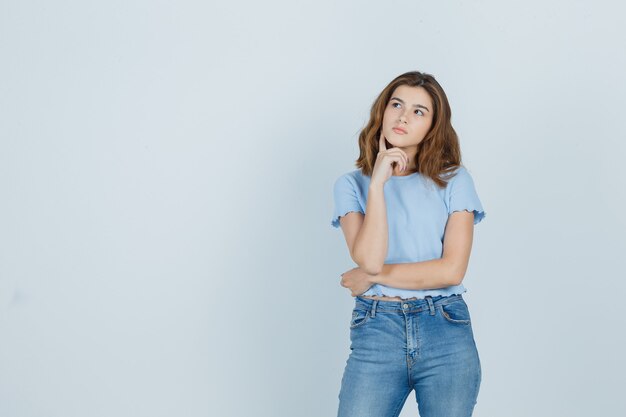 Jovem garota em t-shirt, jeans, segurando o dedo no queixo e olhando pensativa, vista frontal.