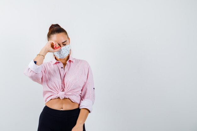 Jovem garota esfregando os olhos com o punho enquanto chorava de blusa rosa, calça preta, máscara e parecendo lúgubre, vista frontal.