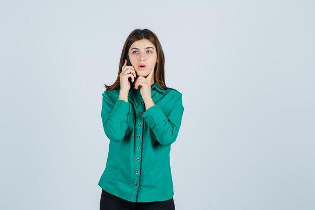 Jovem garota falando ao telefone, colocando o dedo indicador sob o queixo na blusa verde, calça preta e parecendo surpresa. vista frontal.