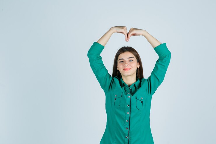 Foto grátis jovem garota fazendo formato de coração com as mãos acima da cabeça na blusa verde, calça preta e olhando alegre, vista frontal.