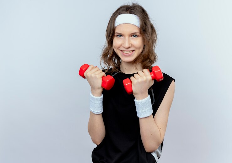 Foto grátis jovem garota fitness em roupa esportiva preta com bandana, malhando com halteres e um sorriso no rosto, em pé sobre uma parede branca