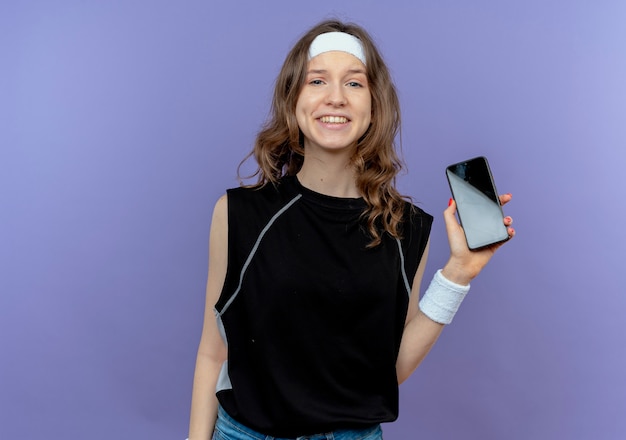 Jovem garota fitness em roupa esportiva preta com fita para a cabeça mostrando smartphone sorrindo alegremente em pé sobre uma parede azul