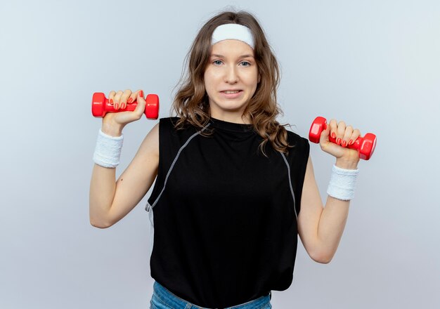 Jovem garota fitness em roupa esportiva preta com tiara, malhando com halteres confusa em pé sobre uma parede branca