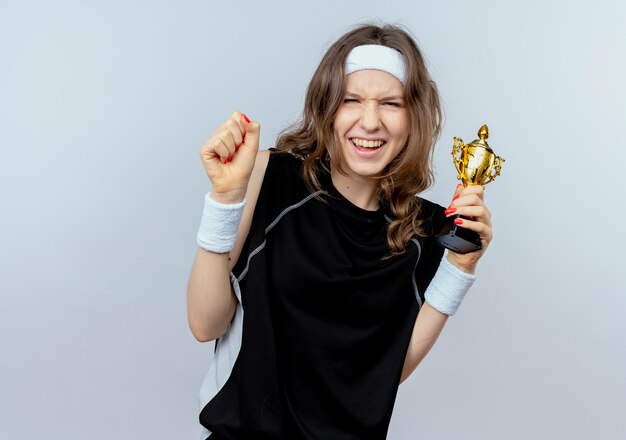 Jovem garota fitness em roupa esportiva preta com tiara segurando um troféu, cerrando o punho, feliz e animada em pé sobre uma parede branca