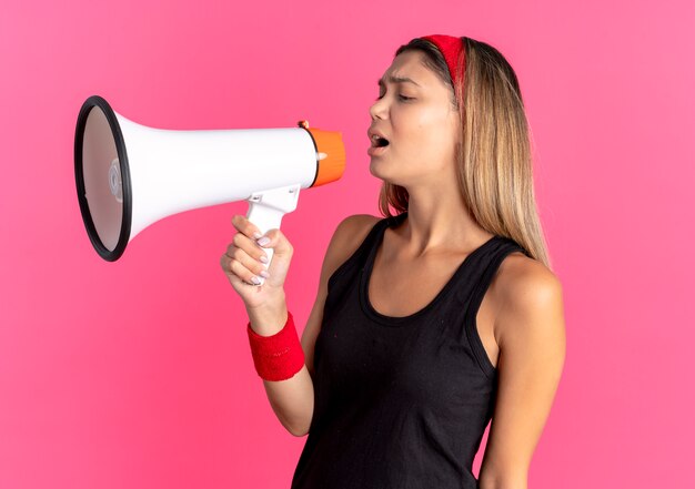 Jovem garota fitness em roupa esportiva preta e bandana vermelha gritando para o megafone em pé sobre a parede rosa