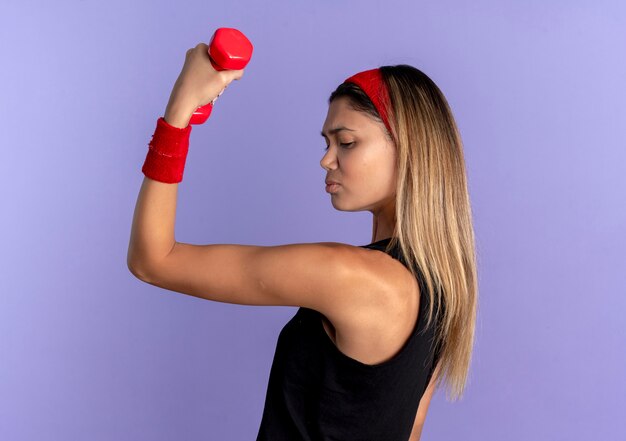 Jovem garota fitness em roupa esportiva preta e bandana vermelha, malhando com halteres mostrando bíceps parecendo confiante sobre o azul