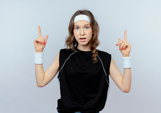Jovem garota fitness em roupas esportivas pretas com fita para a cabeça confusa, apontando com os dedos em pé sobre uma parede branca
