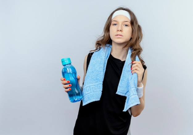 Jovem garota fitness em roupas esportivas pretas com fita para a cabeça e toalha em volta do pescoço segurando uma garrafa de água com cara séria em pé sobre uma parede branca