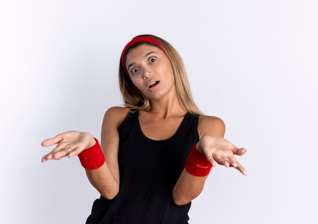 Foto grátis jovem garota fitness em roupas esportivas pretas e bandana vermelha, confusa com os braços abertos, tendo dúvidas em pé sobre uma parede branca