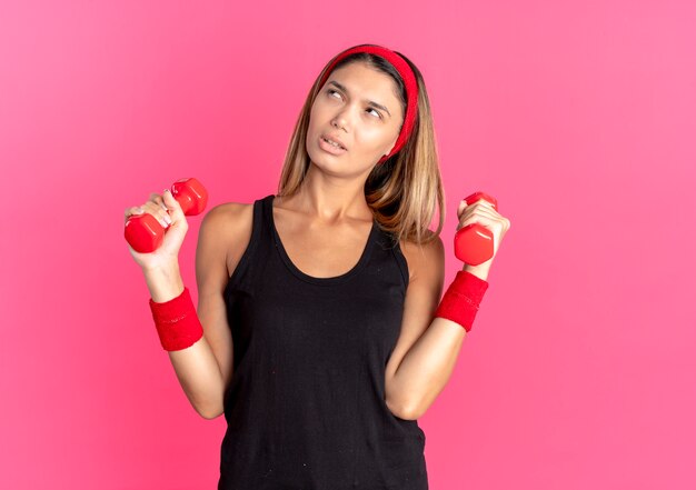 Jovem garota fitness em roupas esportivas pretas e bandana vermelha, malhando com halteres, olhando para o lado perplexa em pé sobre a parede rosa