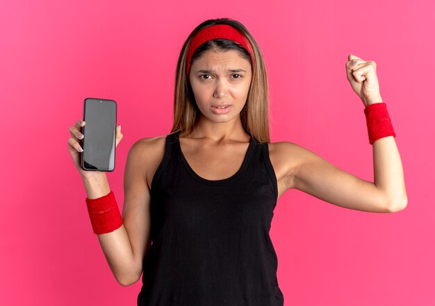 Jovem garota fitness em roupas esportivas pretas e bandana vermelha, mostrando o punho cerrado do smartphone com cara de raiva em pé sobre a parede rosa