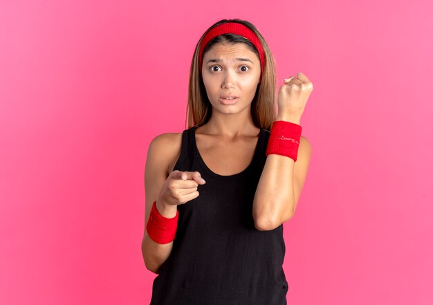 Jovem garota fitness em roupas esportivas pretas e bandana vermelha, parecendo confusa, o punho cerrado apontando com o dedo para a câmera sobre o rosa