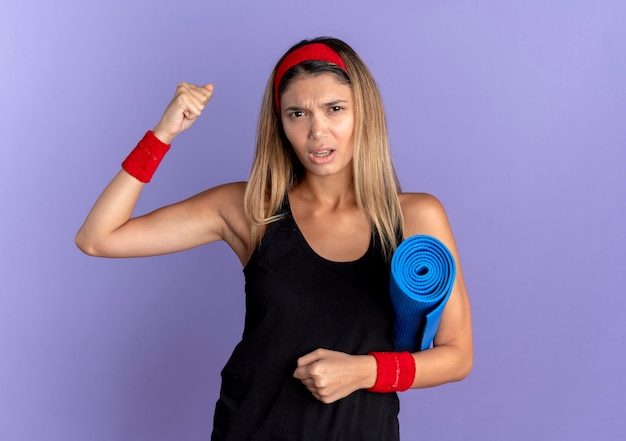 Jovem garota fitness em roupas esportivas pretas e bandana vermelha segurando um tapete de ioga, levantando o punho com uma cara zangada em pé sobre a parede azul