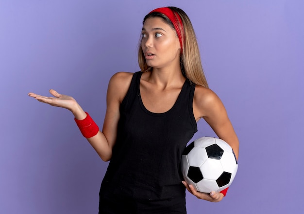 Jovem garota fitness em roupas esportivas pretas e bandana vermelha, segurando uma bola de futebol, apresentando spmething com o braço da mão preocupada em pé sobre a parede azul