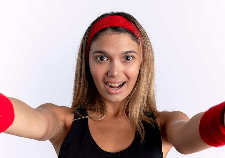 Foto grátis jovem garota fitness em roupas esportivas pretas e bandana vermelha, sorrindo alegremente em pé sobre uma parede branca