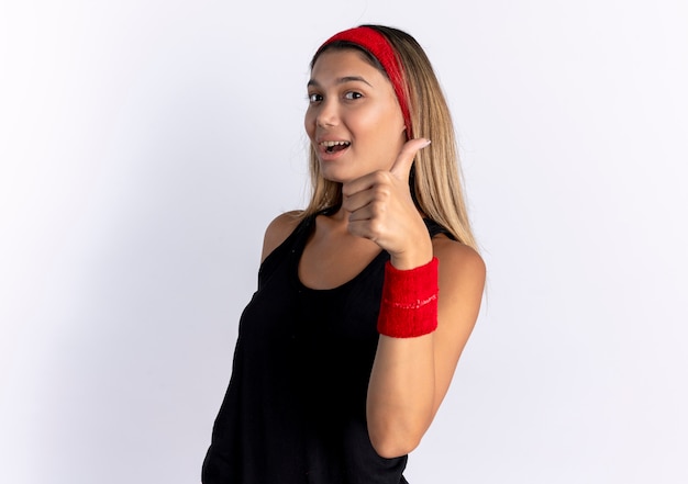 Jovem garota fitness em roupas esportivas pretas e bandana vermelha, sorrindo, mostrando os polegares em pé sobre uma parede branca