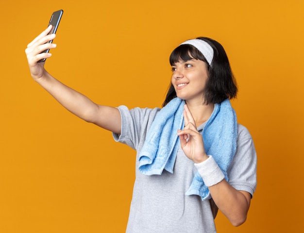 Jovem garota fitness usando bandana com toalha em volta do pescoço fazendo selfie usando smartphone, sorrindo, mostrando o sinal v em pé sobre fundo laranja