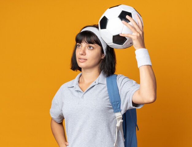 Jovem garota fitness usando bandana e mochila segurando uma bola de futebol, olhando para o lado intrigada