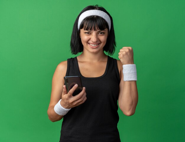 Jovem garota fitness usando bandana e segurando o smartphone com o punho cerrado, feliz e animada olhando para a câmera