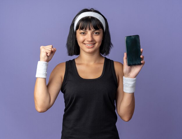 Jovem garota fitness usando bandana e segurando o smartphone com o punho cerrado, feliz e animada olhando para a câmera