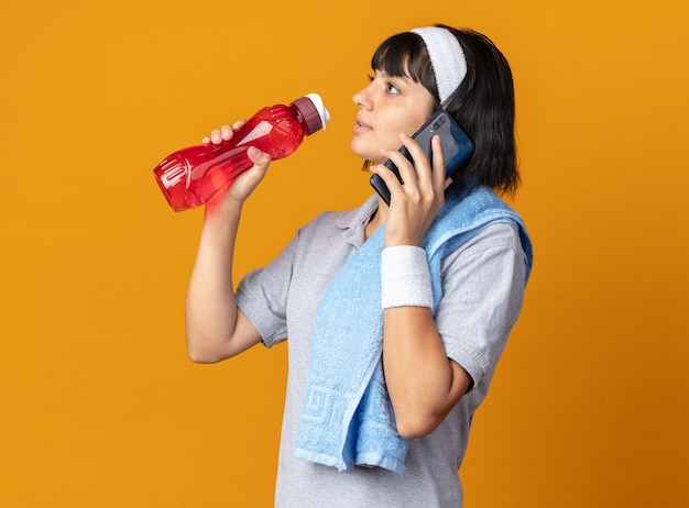 Jovem garota fitness usando bandana e toalha no ombro, segurando uma garrafa de água, parecendo confiante enquanto fala no celular