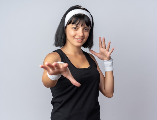 Jovem garota fitness usando bandana, olhando para a câmera, gesticulando com as mãos sorrindo alegremente em pé sobre um fundo branco