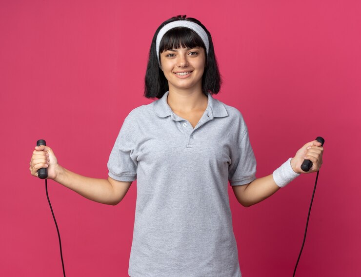 Foto grátis jovem garota fitness usando bandana segurando corda de pular, feliz e alegre, olhando para a câmera, sorrindo em pé sobre um fundo rosa