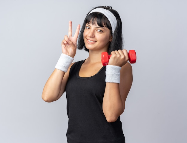 Jovem garota fitness usando bandana segurando halteres, fazendo exercícios, olhando para a câmera, sorrindo e mostrando o sinal-V em pé sobre um fundo branco