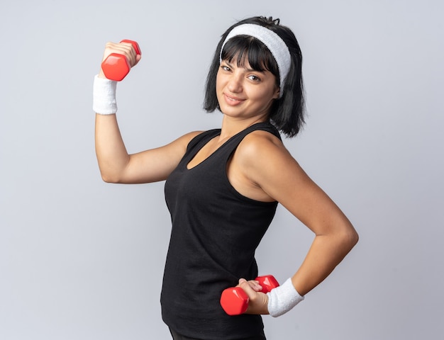 Jovem garota fitness usando bandana segurando halteres, fazendo exercícios, parecendo confiante em pé sobre o branco