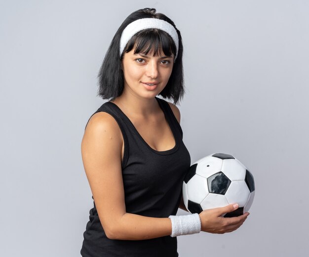 Jovem garota fitness usando bandana segurando uma bola de futebol, olhando para a câmera, sorrindo confiante em pé sobre o branco