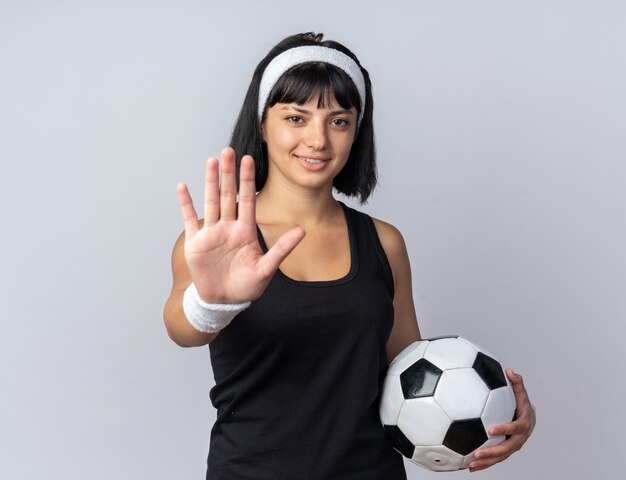 Jovem garota fitness usando bandana segurando uma bola de futebol, olhando para a câmera, sorrindo, fazendo gesto de parada com a mão em pé sobre o branco