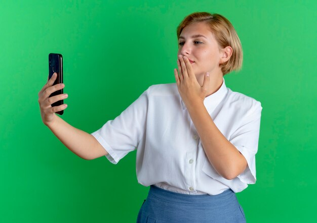 Jovem garota loira russa satisfeita segurando e olhando para o telefone mandando beijo com a mão isolada no fundo verde com espaço de cópia