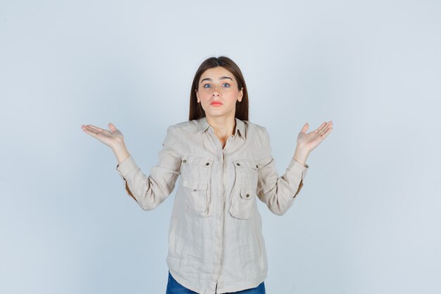 Jovem garota mostrando um gesto impotente em uma camisa bege, jeans e parecendo perplexo, vista frontal.