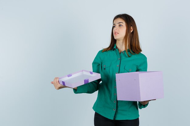 Jovem garota na blusa verde, calça preta abrindo a caixa de presente, olhando para longe e olhando com foco, vista frontal.